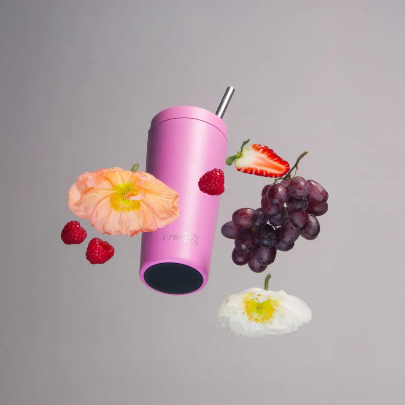 Pink tumbler with a straw surrounded by fruits and flowers on a gray background