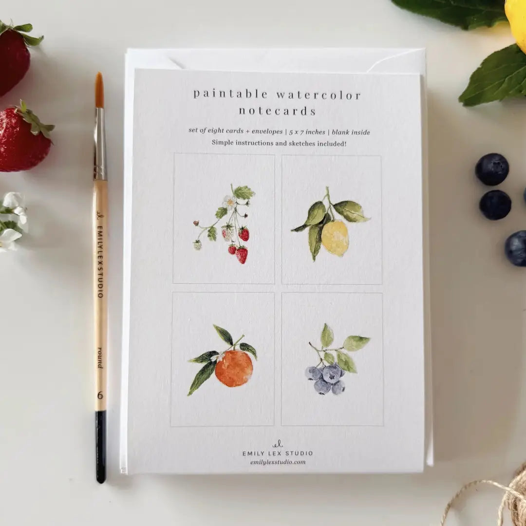 Set of paintable watercolor notecards with illustrations of fruits and flowers, accompanied by a paintbrush and berries on a white surface.