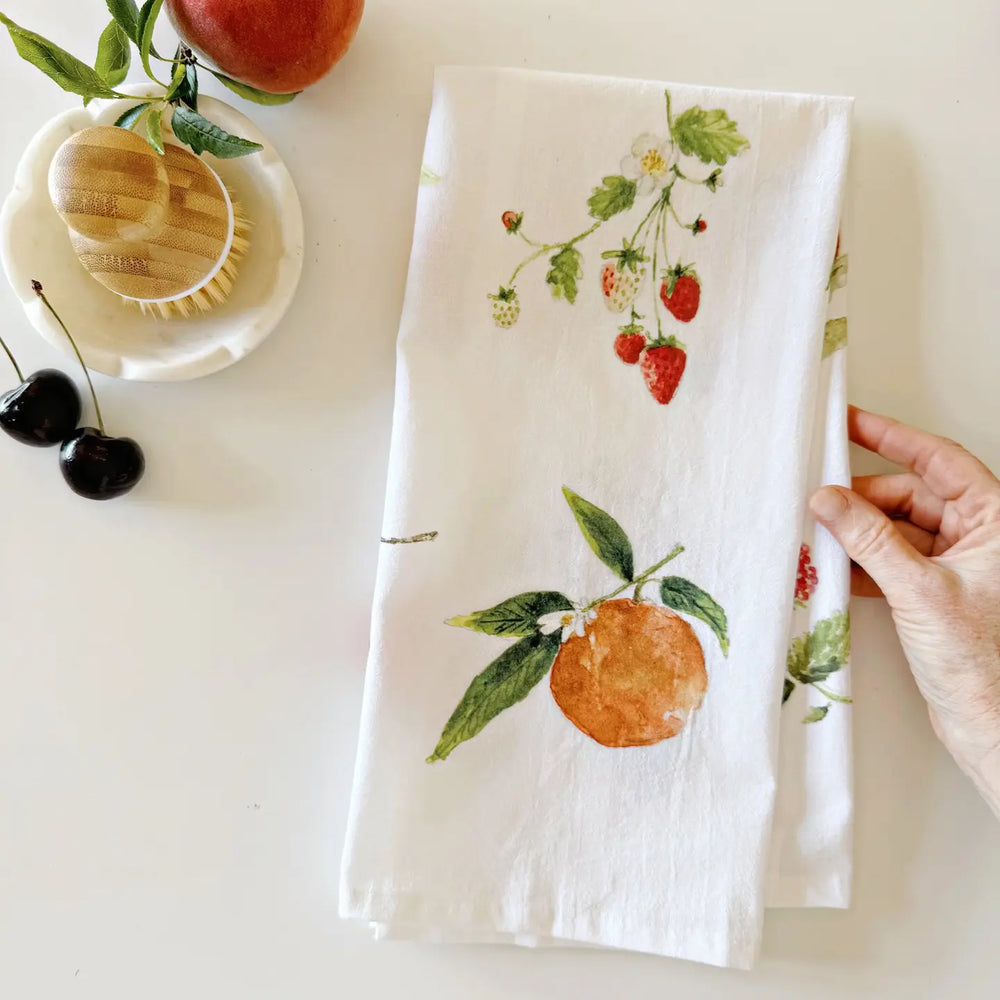 White towel with fruit and strawberry design held by a hand on a white surface with apples and cherries.