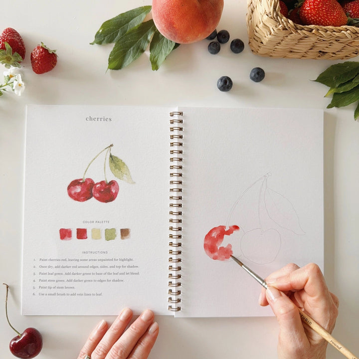 Open notebook with a drawing of cherries, surrounded by real fruits on a light surface.