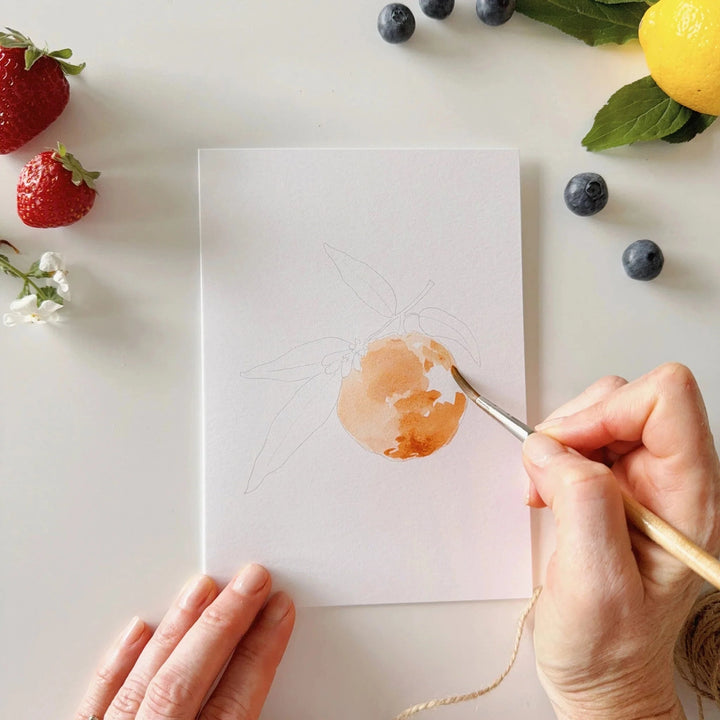 Person painting a watercolor of a strawberry on a white sheet with fruits and flowers around.