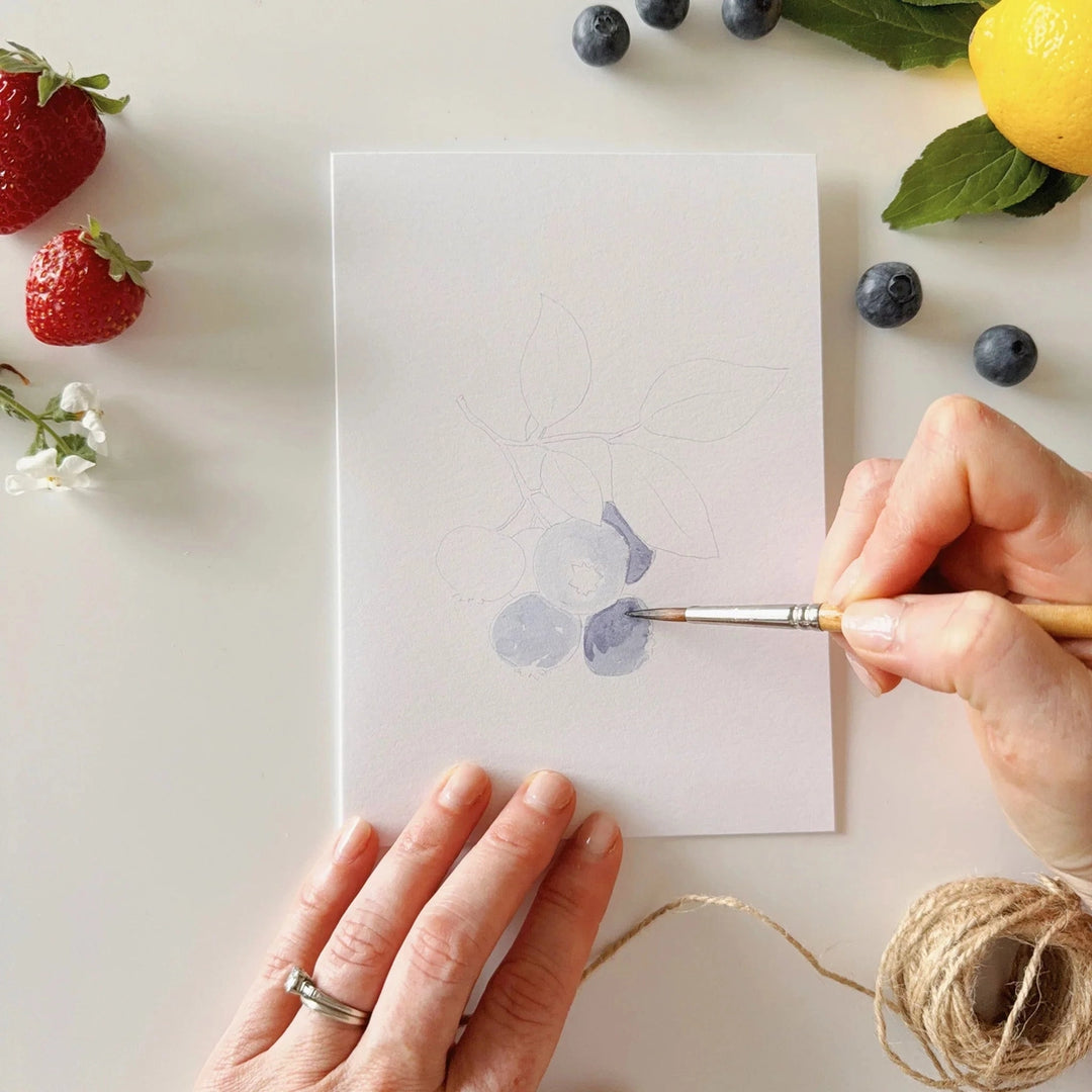 Person painting blueberries on a card with fruits and flowers around