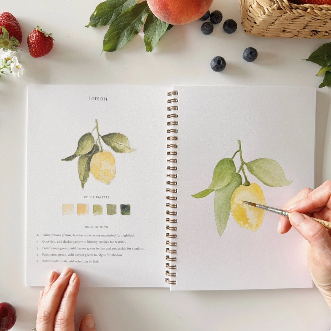 Open book with lemon illustrations on a table with fruits and a hand holding a paintbrush.