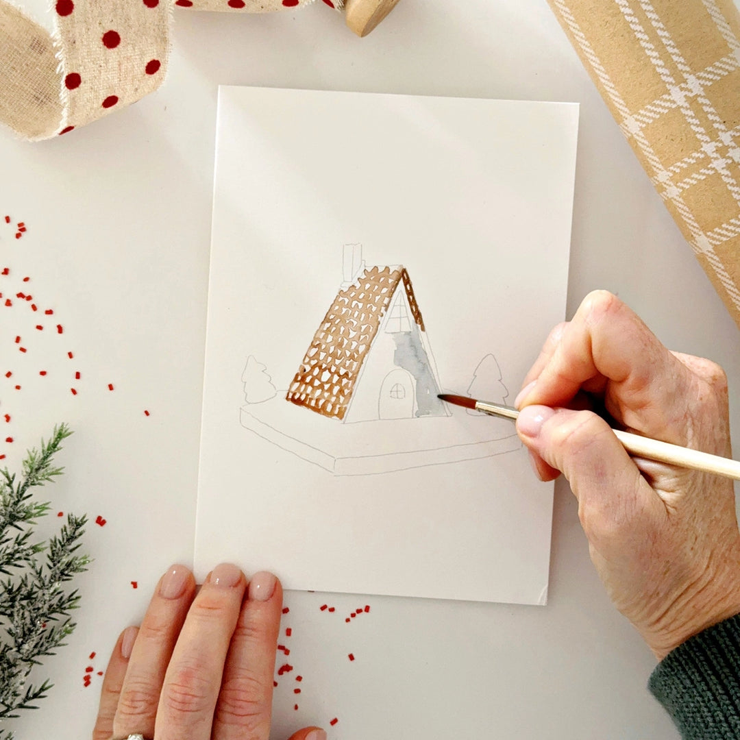 Person painting a small house on a card with decorative elements around