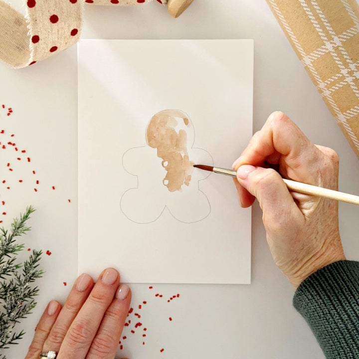 Person painting a gingerbread man on a card with Christmas decorations around