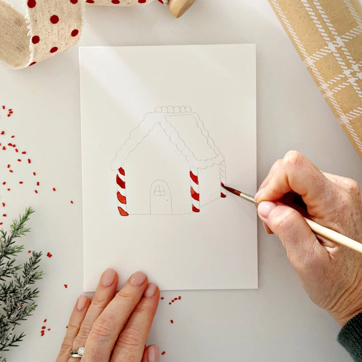 Person painting a gingerbread house design on a card with a brush.