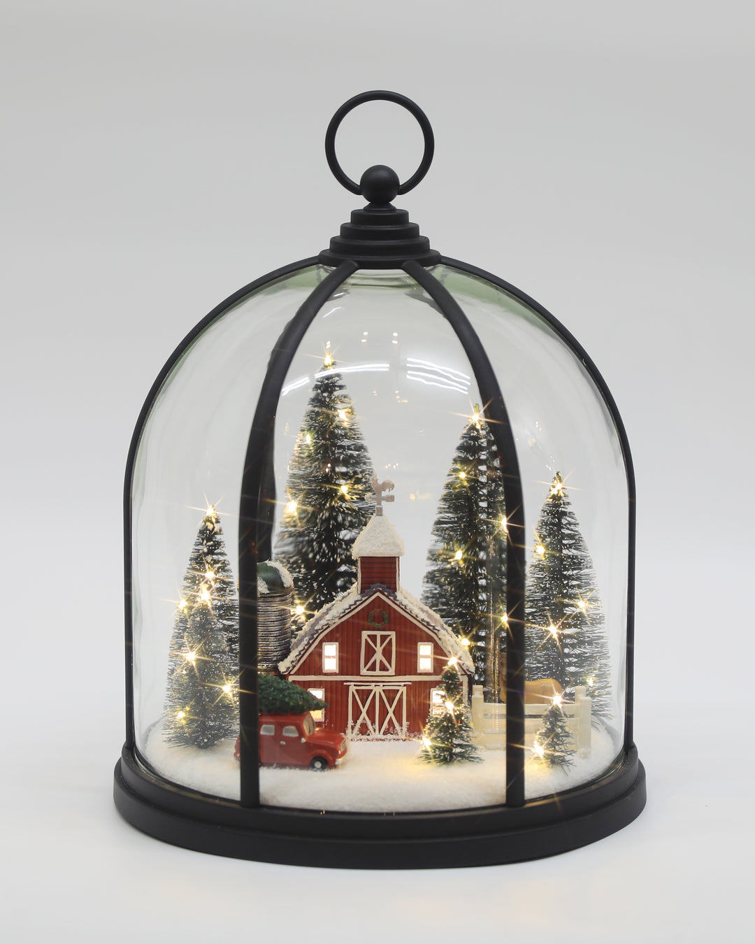 A miniature winter village scene inside a clear glass cloche with a black metal frame and top loop. The scene includes a red barn, small decorated trees, warm white string lights, and a red truck, all displayed on a light gray background.