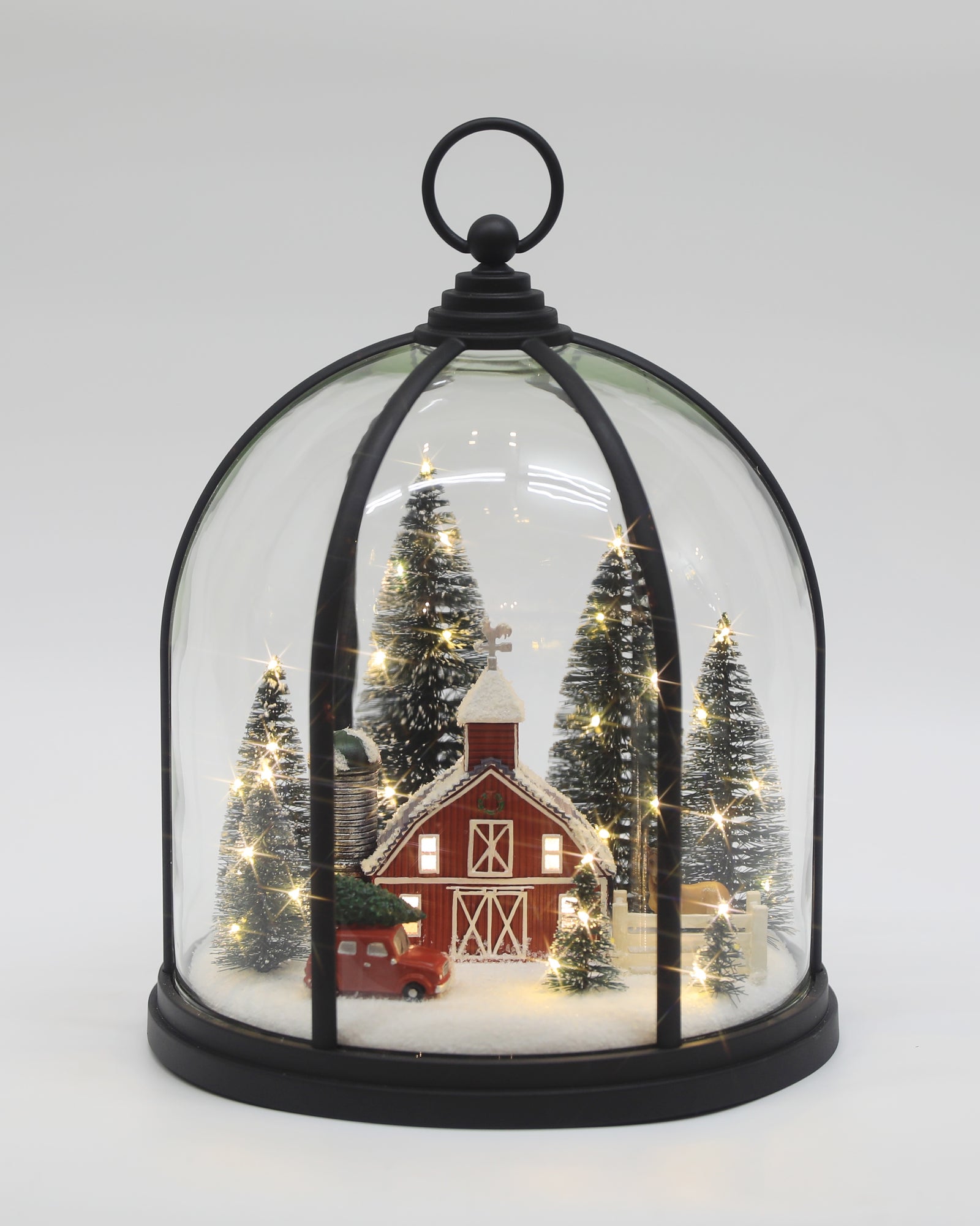 A miniature winter village scene inside a clear glass cloche with a black metal frame and top loop. The scene includes a red barn, small decorated trees, warm white string lights, and a red truck, all displayed on a light gray background.