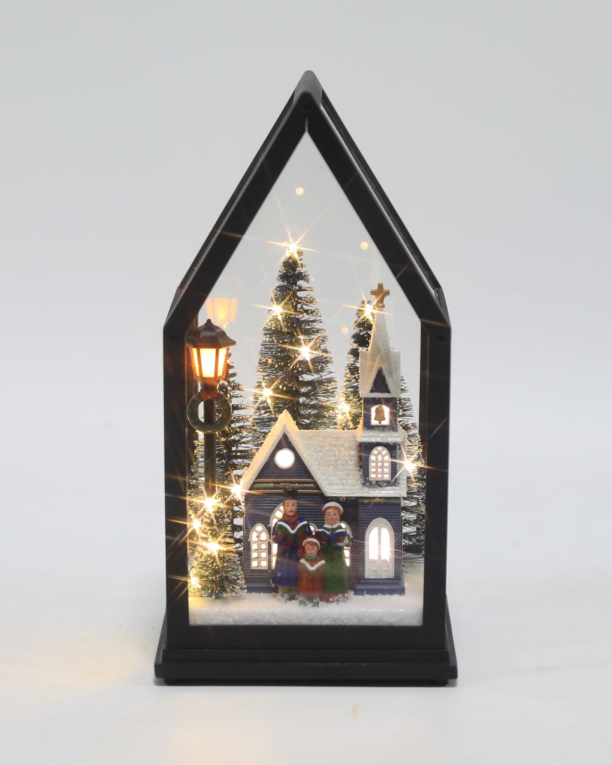 A miniature Christmas village diorama inside a black house-shaped frame, featuring a lit church, decorated trees, a street lamp, and small singing figures in the snow, photographed against a plain light gray background.