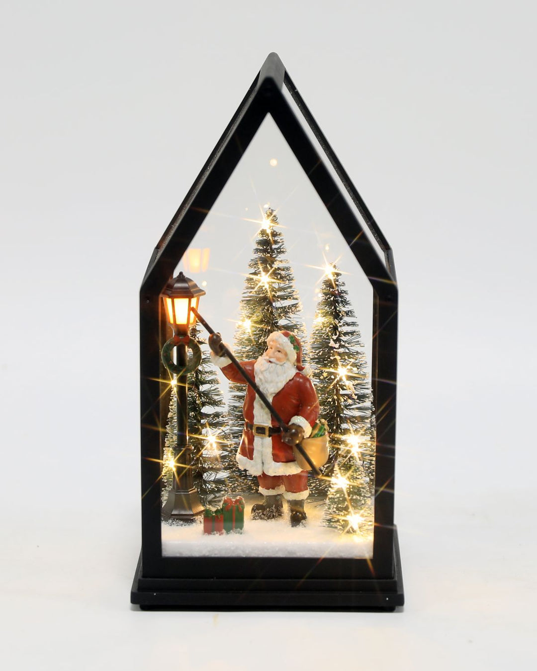 A house-shaped black-framed Christmas table lamp with a Santa Claus scene inside, featuring lit Christmas trees, a street lamp, and warm white fairy lights on a white background.