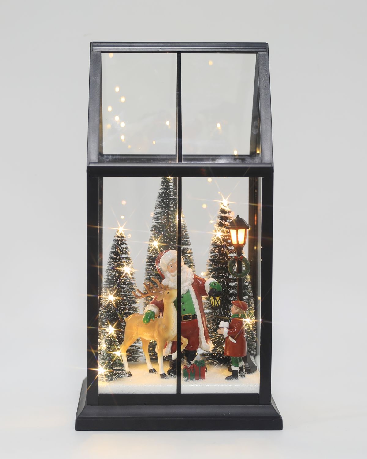 A festive Christmas window lantern decoration with a black frame and warm white lights. Inside, Santa Claus, a reindeer, a chimney sweep, and snow-covered Christmas trees are illuminated against a light gray background.