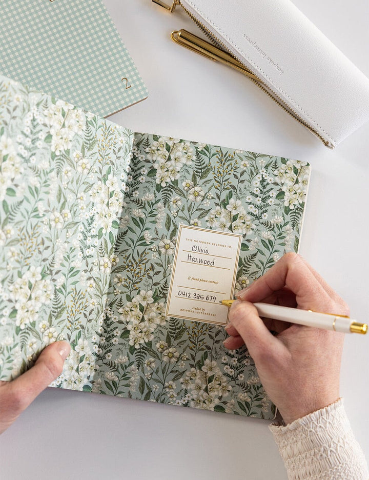 Floral notebook with a hand writing on it, surrounded by other notebooks.