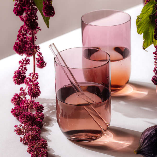 Two pink glasses with a straw on a white surface with purple flowers and green leaves.