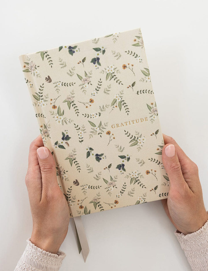 Hands holding a notebook with floral pattern and 'Gratitude' text on a white background