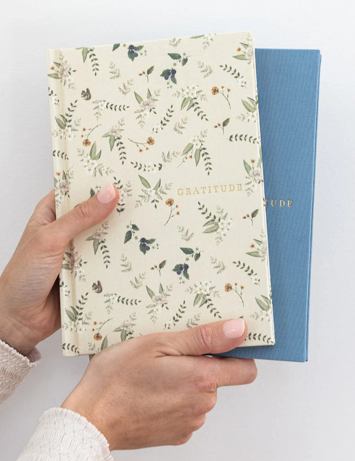 Hand holding a notebook with floral pattern and 'Gratitude' text on a white background