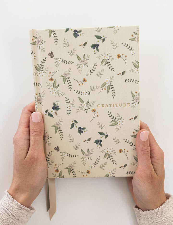 Notebook titled 'Gratitude' with floral design held by hands on a white background
