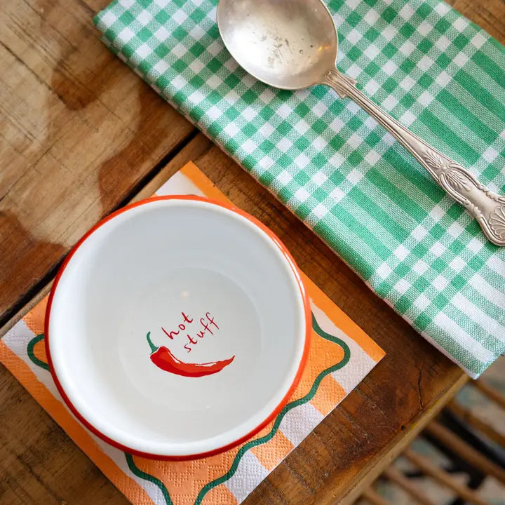 White bowl with red chili pepper design and 'hot stuff' text on a wooden table with a green checkered cloth and silver spoon.