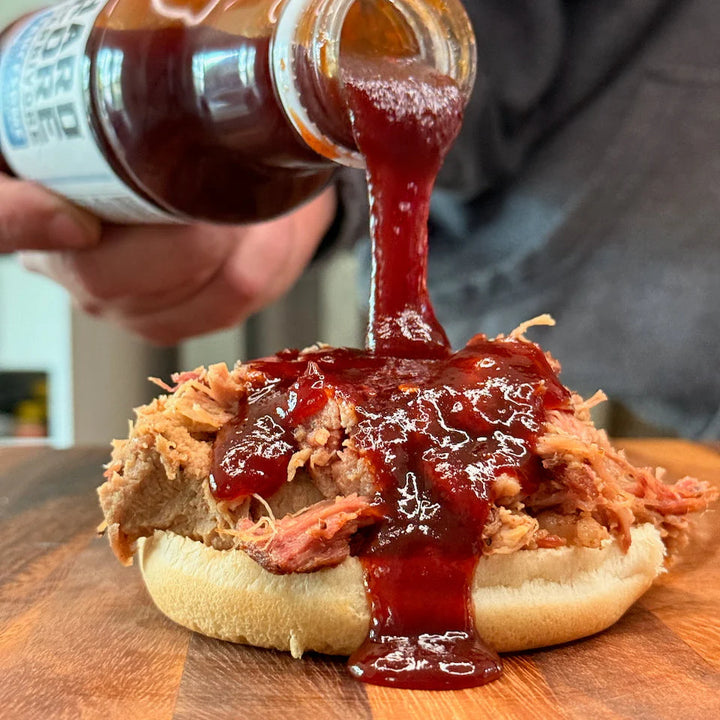 Barbecue sauce being poured onto a pulled pork sandwich on a wooden cutting board.
