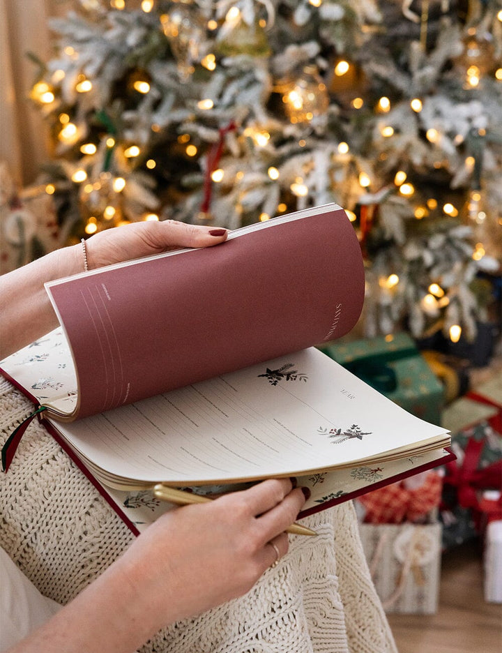 Person holding a red notebook in front of a decorated Christmas tree