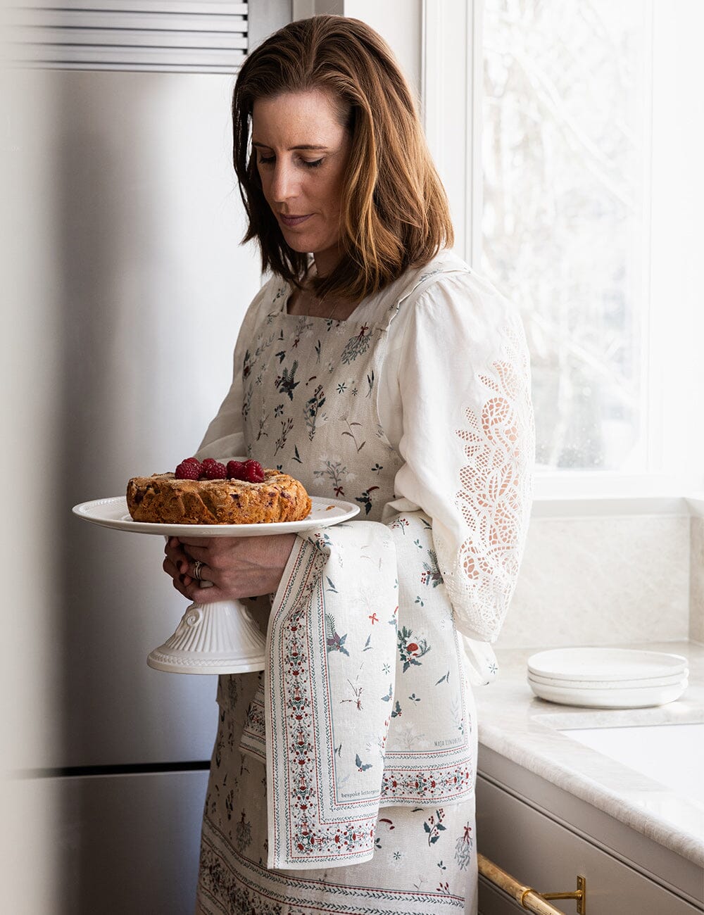 Someone holding a christmas cake with a linen tea towel over her arm and wearing a matching christmas apron