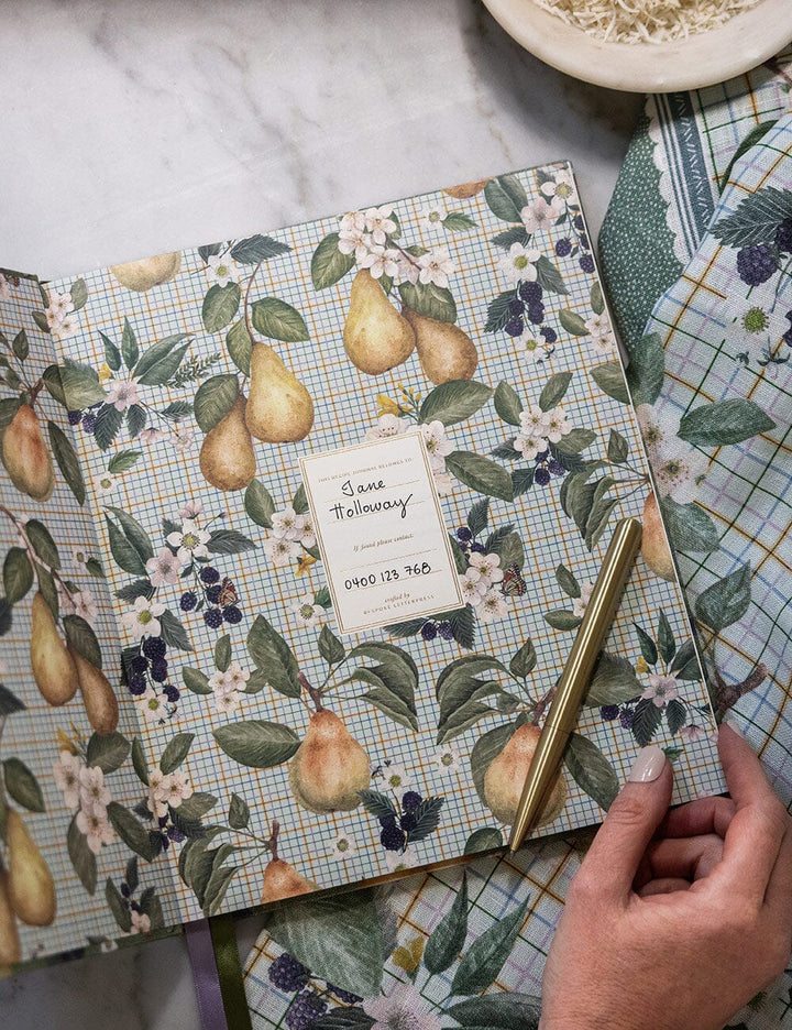 Notebook with pear and floral design on cover, held by a hand with a marble background.