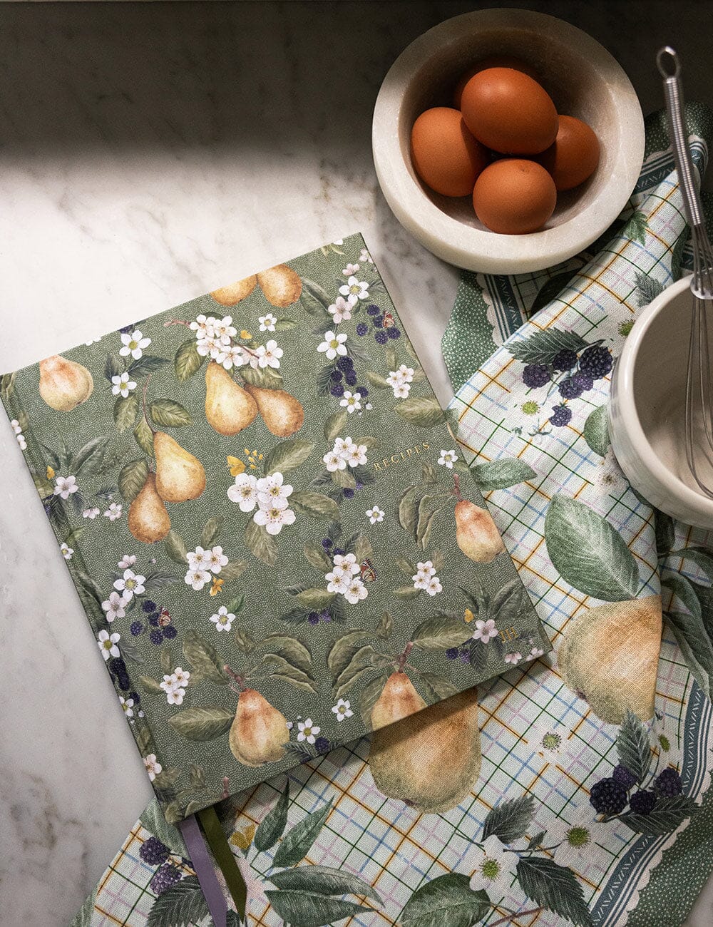 Notebook with floral and fruit design on a kitchen counter with eggs and utensils.