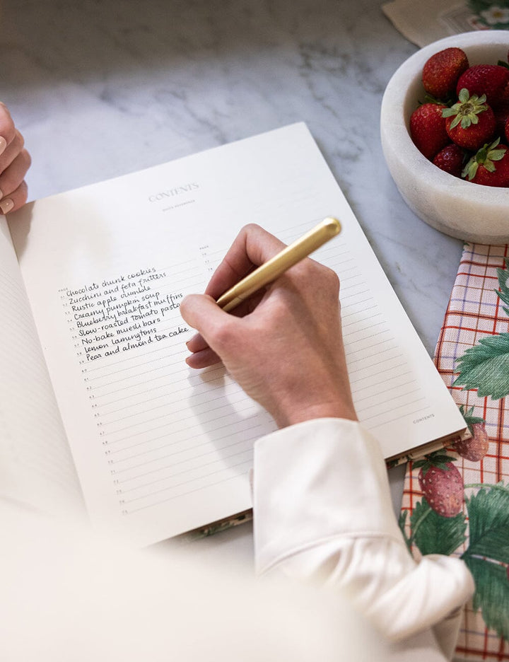 Person writing in a notebook with a pen, next to a bowl of strawberries on a marble surface.