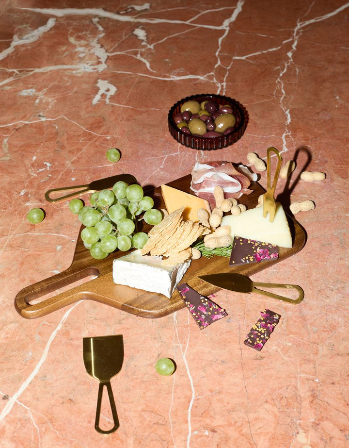 Cheese board with cheese and crackers on it surrounded by gold cheese knives on a pink marble background