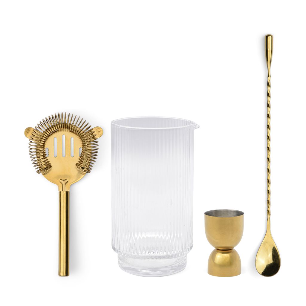 Set of bar tools including a gold strainer, clear glass, gold jigger, and gold spoon on a white background.