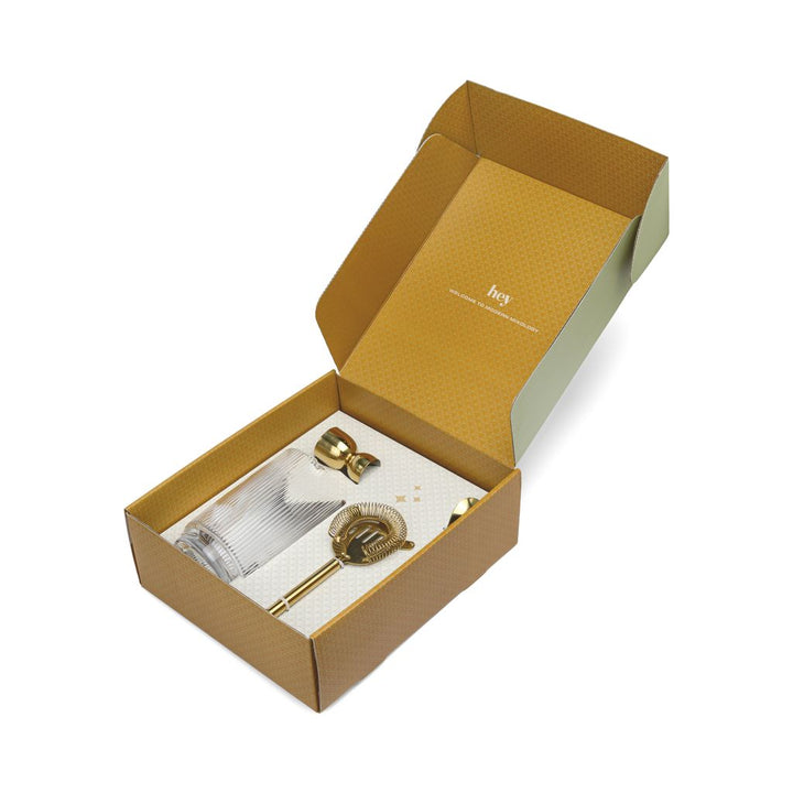 Open cardboard box with gold and glass cocktail accessories on a white background