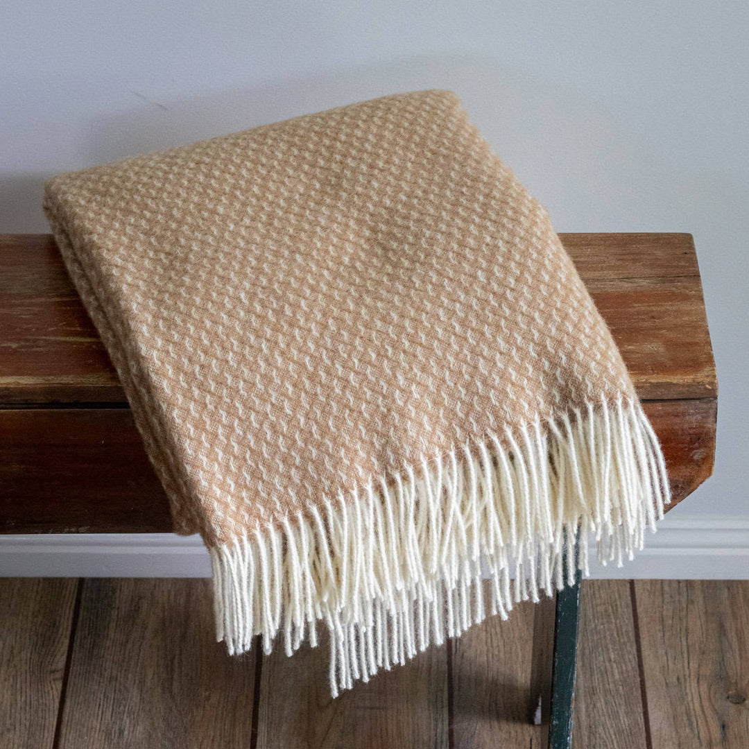 Beige woven throw blanket with white fringe on a wooden bench against a plain wall.