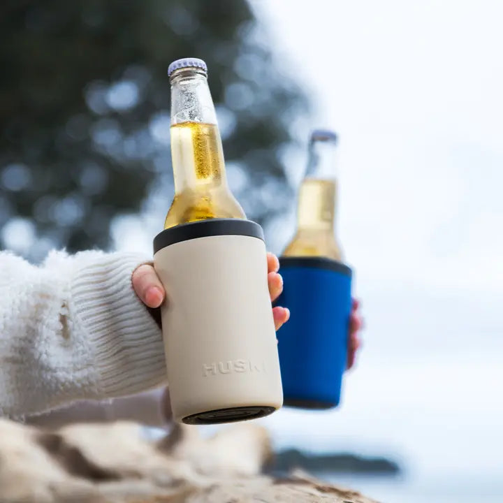 Two bottles of beer held by a person wearing a white sweater, one in a beige cooler and the other in a blue cooler.