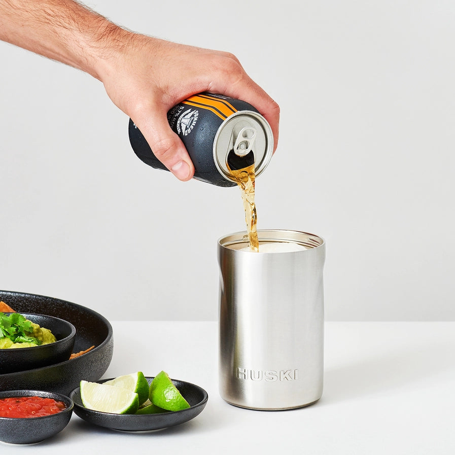 Person pouring beer from a can into a HUSKIS tumbler with food items on a white background
