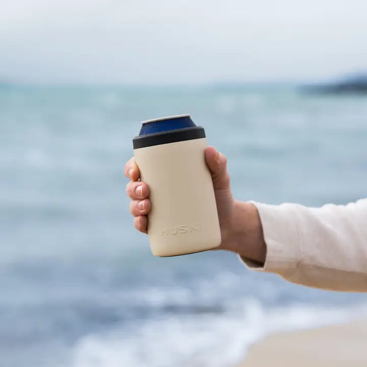 Hand holding a beige can cooler with a blue label against a blurred ocean background