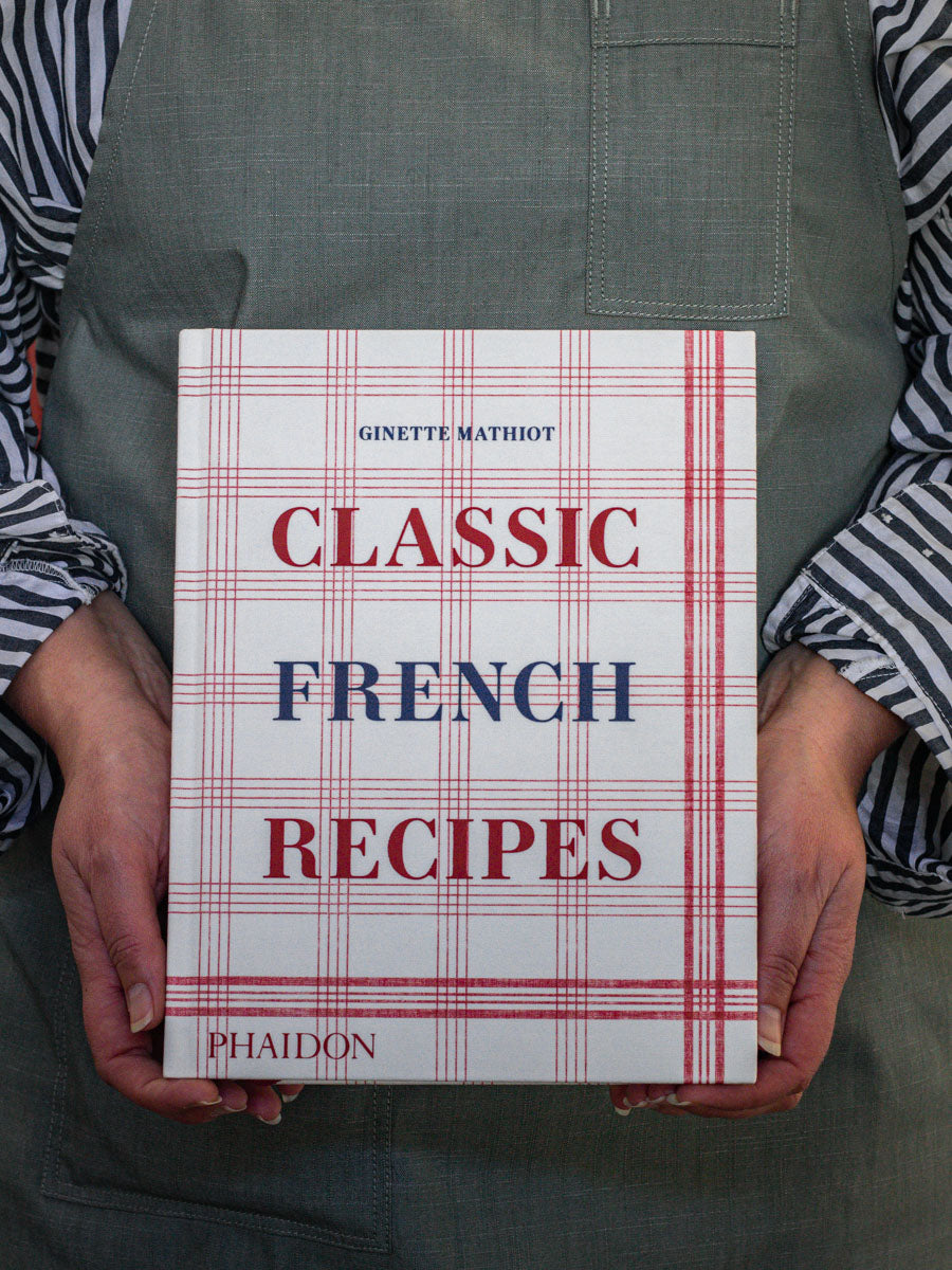 A person holding a book titled 'Classic French Recipes' by Ginette Mathiot, with a red and white checkered design on the cover.