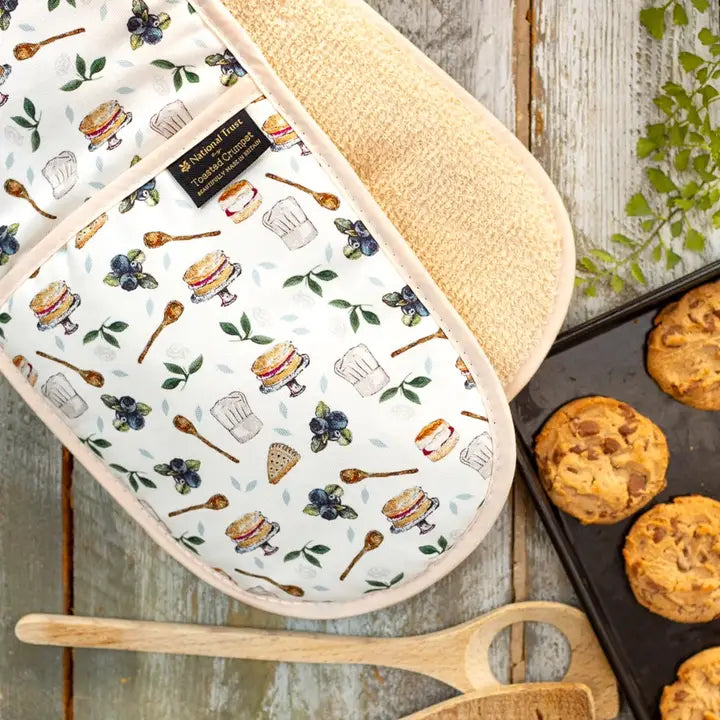 Double oven glove with food-themed pattern on a wooden surface with cookies and a spatula.
