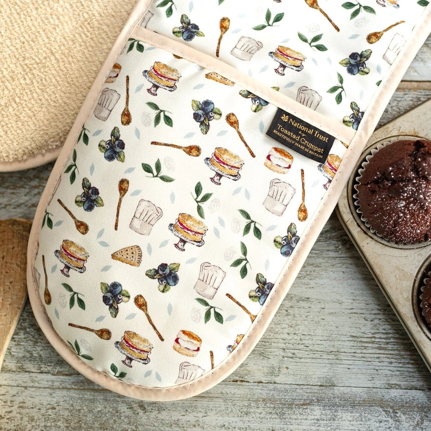 Oven mitt with baking-themed design on a wooden surface with a muffin tin.