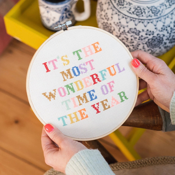 Embroidery hoop with colorful text held by a person, with a blurred background of a kitchen setting.
