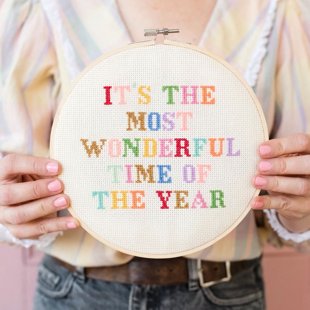 Person holding a cross-stitched hoop with colorful text 'IT'S THE MOST WONDERFUL TIME OF THE YEAR'.