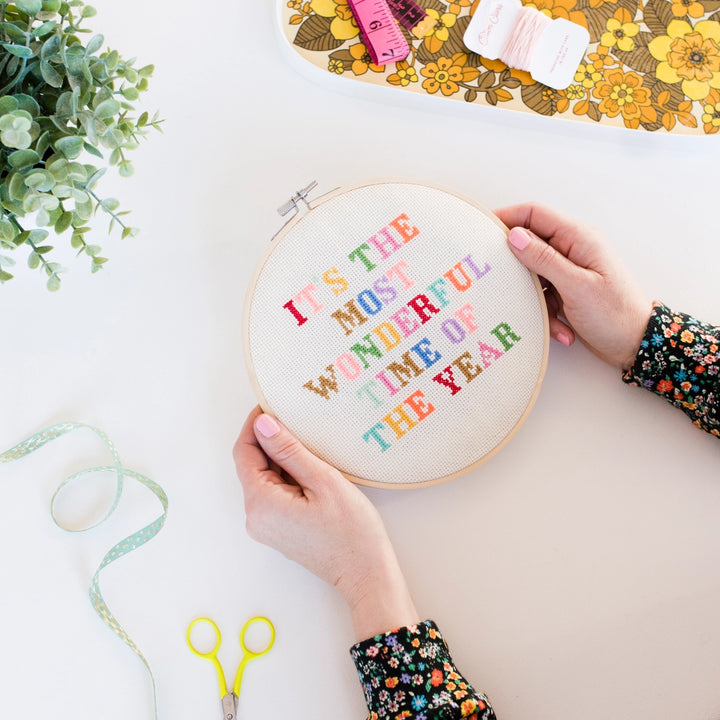 Embroidery hoop with colorful text held by a person on a table with crafting supplies.