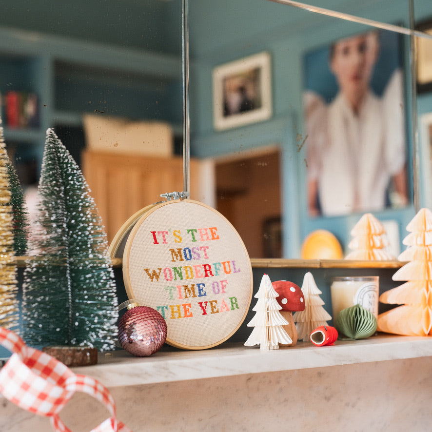 Decorative shelf with Christmas trees, ornaments, and a cross stitch reading 'It's the Most Wonderful Time of the Year'.