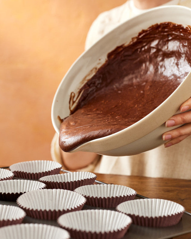 Person pouring chocolate batter into cupcake liners on a wooden table.