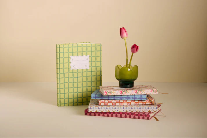 Stack of patterned books with a green vase containing pink flowers on a beige surface.