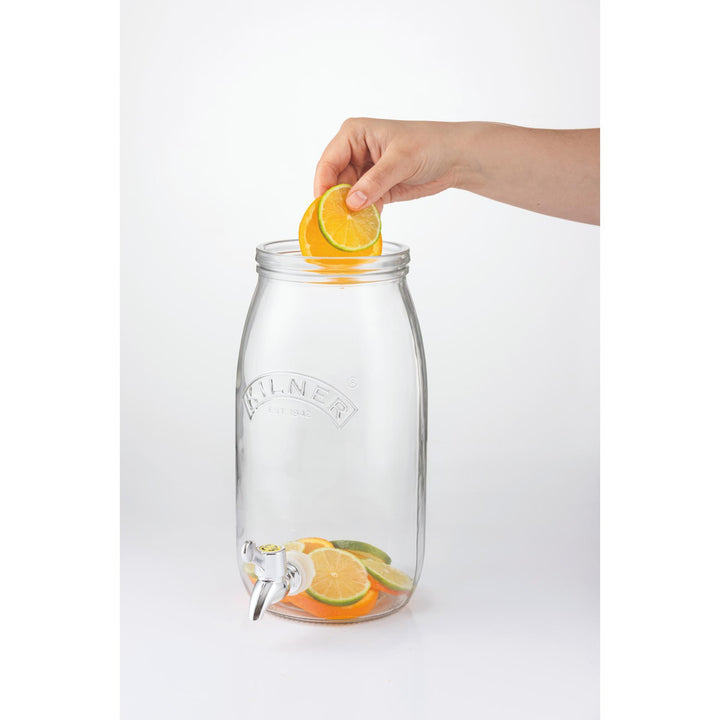 Clear glass drink dispenser with lemons and limes, hand placing a lemon slice on top.