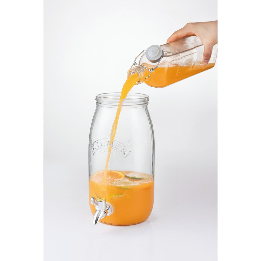 Orange juice being poured from a carafe into a glass jar with a spigot on a white background