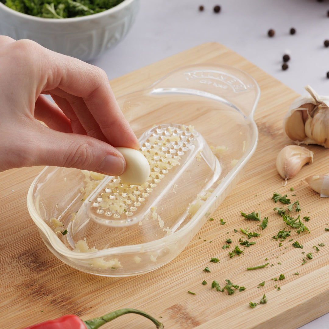 Hand using a grater on a wooden cutting board with chopped garlic and herbs.
