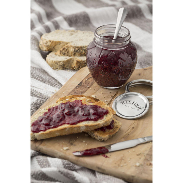 Jar of jam with a spoon, slices of bread with jam, and a wooden board on a textured fabric background.