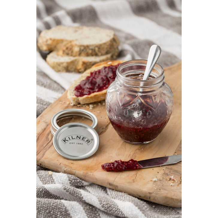 Jar of jam with a spoon, bread slices, and a Kilner jar lid on a wooden board.