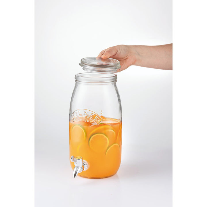 Glass drink dispenser with orange liquid and a hand placing a lid on it, on a white background