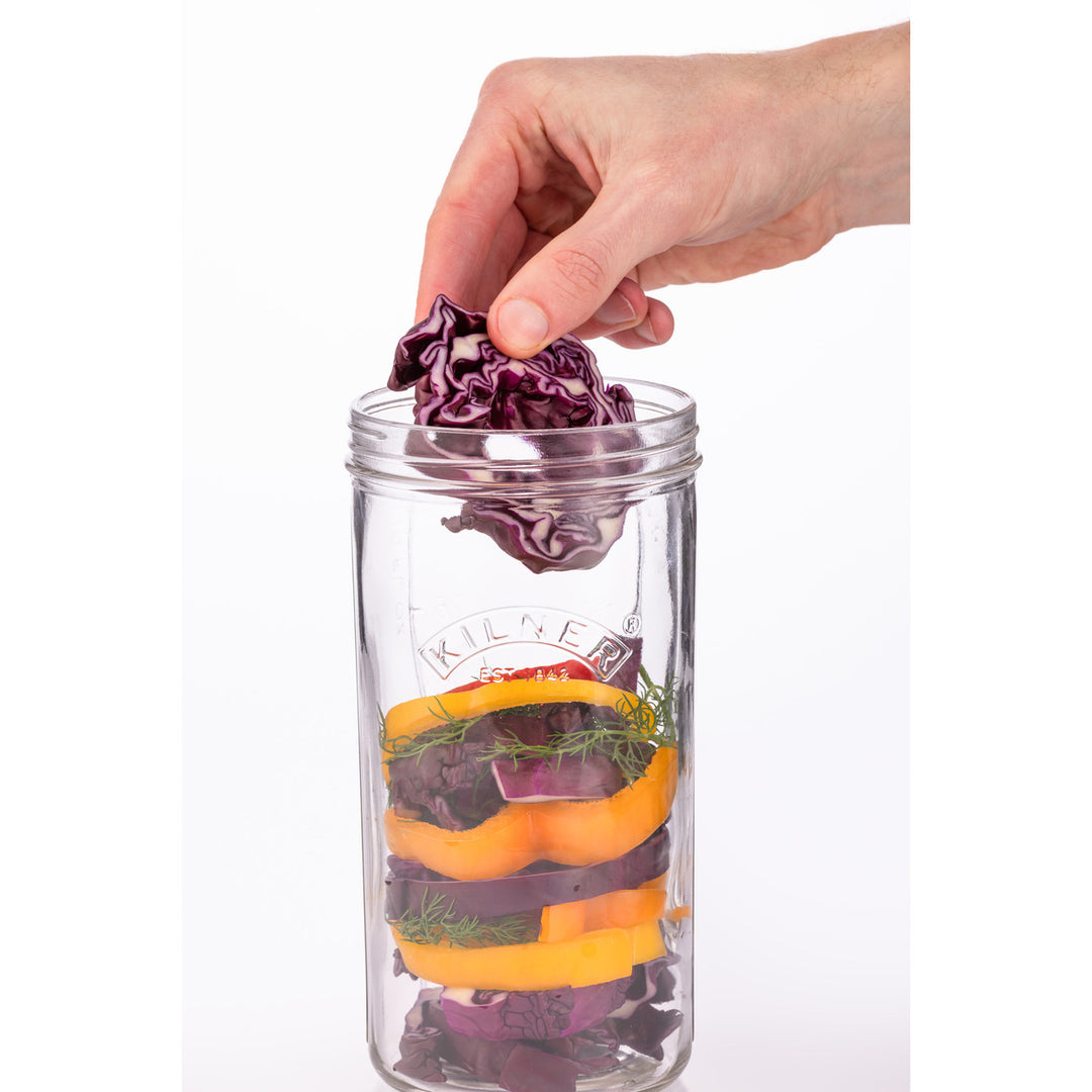 Hand placing a purple cabbage into a clear jar with other vegetables on a white background